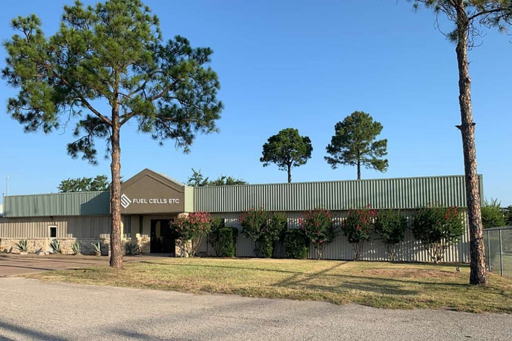 Fuel Cells Etc manufacturing building in Bryan, Texas, behind tall pine trees and flowering shrubs